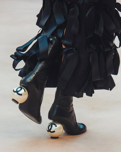 Bottines noires en cuir avec talons ornés de perles siglées Chanel, portées avec une jupe noire à volants.
