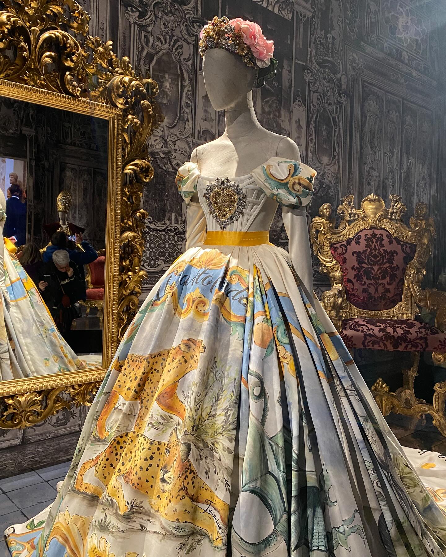 Robe de haute couture exposée sur mannequin, avec jupe illustrée, corset brodé et décor baroque en arrière-plan.
